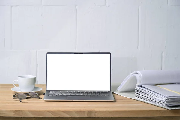 Mock-up of a laptop with blank white screen, file folder and coffee cup on a wooden desk in front of a white painted wall, concept for office and business work, copy space, selected focus