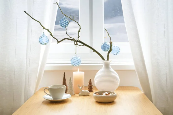 Minimalist holiday decor with blue glass baubles hanging on a bare branch in a vase, candle, small Christmas decoration objects, cookies and coffee on a table by the window, copy space, selected focus