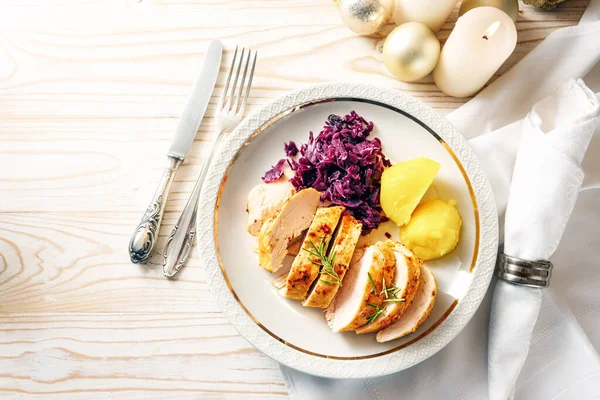 Chicken fillet with red cabbage and potatoes on a white plate, cutlery, napkin and Christmas decoration on a wooden table, holiday dinner, top view from above, copy space, selected focus