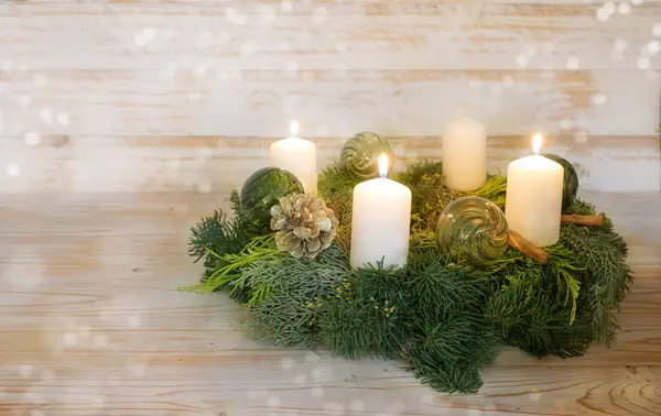 Third advent, natural advent wreath with white candles, tree are lit, green Christmas glass baubles, pine cones and cinnamon, wooden background with snowy bokeh, copy space, selected focus