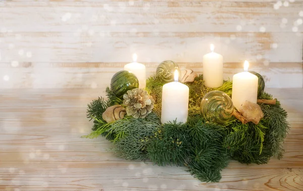 Fourth advent, natural advent wreath with white candles, four are lit, green Christmas glass baubles, pine cones, cinnamon and cookies, wooden background with snowy bokeh, copy space, selected focus