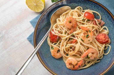 Beyaz ahşap bir masada kaşıkla mavi bir tabakta limon soslu spagetti, karides, domates ve otlar, ev yapımı Akdeniz ürünleri, kopya alanı, yukarıdan üst görünüm, seçilmiş odaklanma
