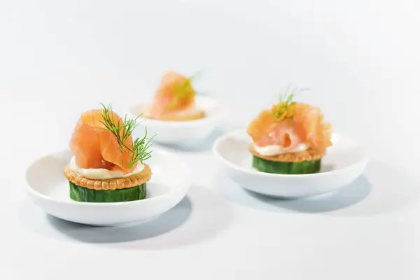 Canapes as party finger food for the cold buffet made from cucumber, crackers, cream cheese, smoked salmon and dill on small white plates, copy space, selected focus, narrow depth of field