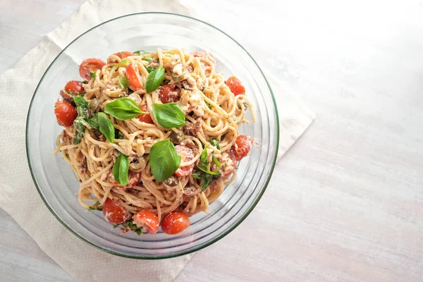Cold spaghetti salad with tomatoes, arugula, mozzarella, olives and a creamy dressing with basil garnish in a glass bowl on a white painted table, top view from above, copy space, selected focus