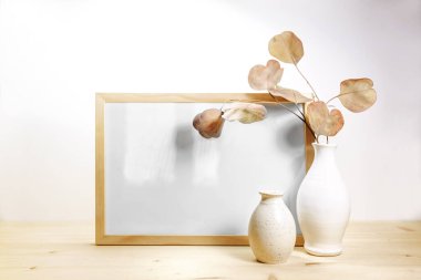Boş resim çerçevesi ve iki seramik vazo, kırmızı yapraklı kuru okaliptüs dalı, açık bej bir duvar, ürün arka planı, kopyalama alanı, seçilmiş odak noktası, ahşap bir masada minimum yaşam