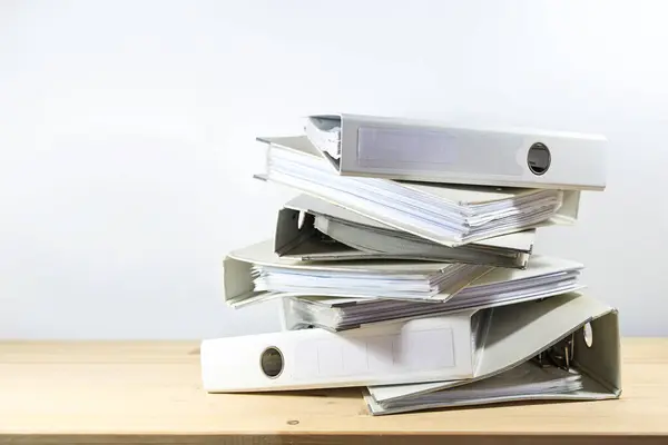 Stack of ring binders on a wooden office desk against a light gray wall, concept of bureaucracy, administration and work overload at the job, copy space, selected focus, narrow depth of field