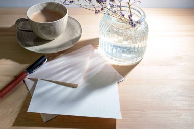 Tebrik kartı ve zarf, doğal geri dönüşüm kağıdı, kalem, kahve fincanı ve cam vazodan sabah tahta bir masada güneş ışığı yansımaları, sıcak bej rengi, kopyalama alanı, seçilmiş odak noktası.