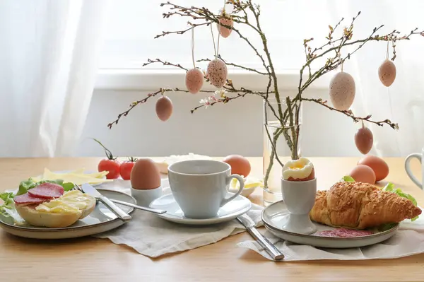 Easter breakfast with egg, bun, coffee, croissant, cheese and sausage, decorated with spring branches and hanging easter eggs in a glass vase on a table at the window, copy space, selected focus