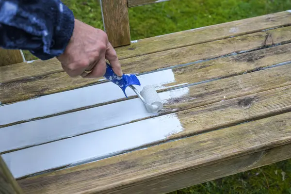 人类正在用漆皮滚筒在户外用白色的防风涂料粉刷一条生机勃勃的木制长椅 为花园季节添置新潮的户外家具 复制空间 选定焦点 图库照片