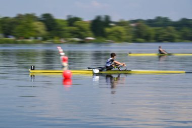 Bir spor teknesindeki tek kürek yarışçısı bitiş çizgisini ilk geçer ve göldeki yelkenli yarışını kazanır, hareket bulanıklığı, kopyalama alanı ve seçilmiş odak noktasıyla döner.
