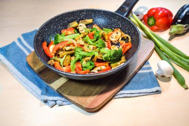 Kızartma tavasında servis edilen karışık sebzeler, dolma biber, brokoli, mantar, patlıcan ve soğan, ahşap kesme tahtasında sağlıklı vejetaryen yemeği ve mutfak havlusu, kopyalama alanı, seçilmiş odak