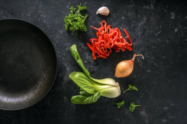 Koyu bir arka planda siyah demir bir tavanın yanında bok choy ya da bak choi, kırmızı dolma biber, soğan, sarımsak ve maydanoz gibi sebzeler, kopya alanı, yukarıdan yüksek açı görünümü, seçilmiş odaklanma açısı