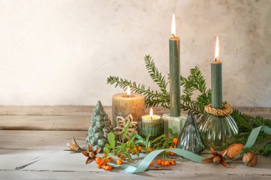 Dört farklı mum, dal, böğürtlen, fındık ve bej kaplama bir duvara karşı kırsal ahşap döşeme, kopyalama alanı ve seçilmiş odaklı Noel süslemesi.