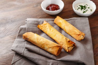 Gri bir peçete ve ahşap bir masa, ev yapımı parmak yiyecekler, fotokopi alanı, seçilmiş odak noktası, dar bir alan derinliği olan üç çıtır fileto böreği ya da yumurta rulosu.