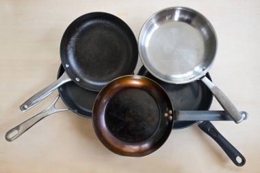 Çeşitli malzemelerden yapılmış kızartma tavalarının seçimi, paslanmaz çelik, dökme demir, karbon çelik ve PTFE yapışmaz kaplama, yukarıdan yüksek açılı görünüm, açık ahşap zemin, seçilmiş odaklanma