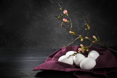 Mor peçetedeki beyaz yumurtalar ve koyu gri arkaplanda pembe tomurcuklu bıldırcın dalı, koyu Paskalya süslemesindeki ışık, kopyalama alanı, seçilmiş odak, dar alan derinliği
