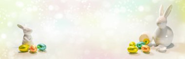 İki beyaz seramik Paskalya tavşanı solda ve sağda küçük renkli çikolatalı yumurta ile pastel renkli açık arkaplan, geniş panoramik format, kopyalama alanı, seçilmiş odak noktası.