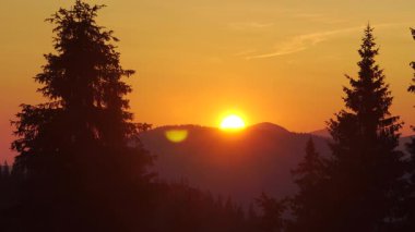 Dağlarda portakal rengi gün batımı ve ön planda çam ağaçları.