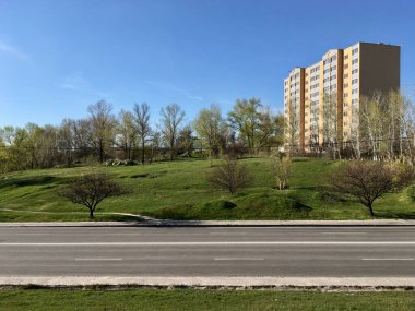 Mavi bulutsuz gökyüzünün altında asfalt iki şeritli yol boyunca büyüyen genç ağaçlarla yeşil tepelerde duran kumlu sarı duvarları olan on katlı bir apartman.