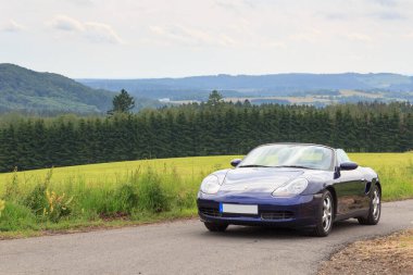 Gerolstein, Almanya - 28 Haziran 2021: Mavi Roadster Porsche Boxster 986 ve Eifel Dağı Panoraması. Araba, Alman otomobil şirketi Porsche tarafından üretilen iki koltuklu bir spor arabadır..