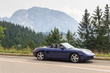 Berchtesgaden, Germany - July 24, 2021: Blue roadster Porsche Boxster 986 with mountain Hoher Goell panorama at Rossfeldpanoramastrae (horse field panoramic motorway). It is a sports car by Porsche.