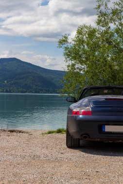 Kochel, Almanya - 29 Temmuz 2023: Mavi Porsche 911 üstü açık otopark Kochelsee Gölü ve Bavyera 'daki dağ alpleri manzarasının yanında.