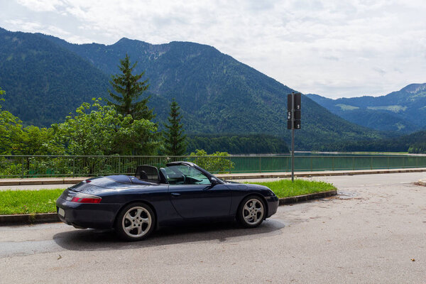Lenggries, Germany - July 29, 2023: Blue Porsche 911 convertible next to Faller-Klamm-Bruecke with lake Sylvenstein Reservoir and mountain alps panorama in Bavaria.