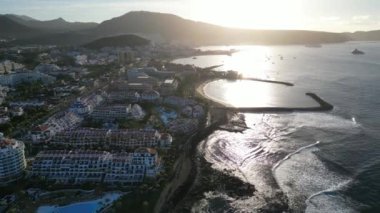 İspanya 'nın Tenerife Kanarya Adası' ndaki Playa de las Americas tatil beldesinde güneş doğarken oteller, gezinti güvertesi, Atlantik Okyanusu ve plajla Los Cristianos 'a doğru uçuş.