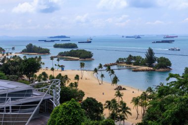 Singapur 'daki Sentosa Adası' nda palmiye ağaçları, adalar ve konteyner gemileriyle Siloso sahili manzaralı.