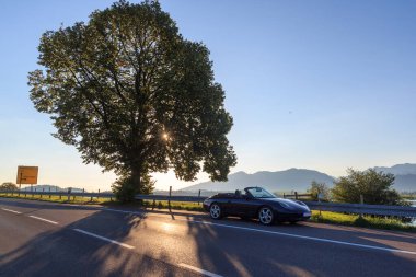 Rieden am Forggensee, Almanya - 10 Ağustos 2024: Bavyera 'da bir ağaç ve dağ Alpleri panoramasının yanında arka plan ışığı ve güneş ışınlarıyla üstü açık mavi Porsche 911