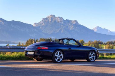 Rieden am Forggensee, Almanya - 10 Ağustos 2024: Blue Porsche 911 Forggensee gölünün yanında üstü açık araba ve Bavyera 'daki dağ alpleri panoraması
