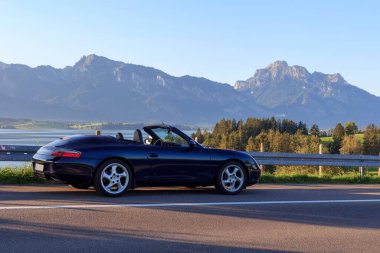 Rieden am Forggensee, Almanya - 10 Ağustos 2024: Blue Porsche 911 Forggensee gölünün yanında üstü açık araba ve Bavyera 'daki dağ alpleri panoraması