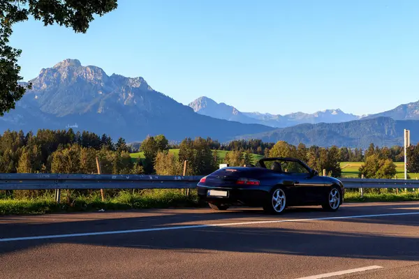 Rieden am Forggensee, Almanya - 10 Ağustos 2024: Blue Porsche 911 Forggensee gölünün yanında üstü açık araba ve Bavyera 'daki dağ alpleri panoraması