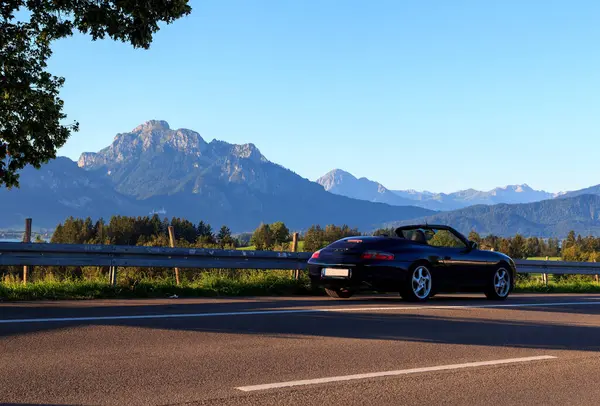 Rieden am Forggensee, Almanya - 10 Ağustos 2024: Blue Porsche 911 Forggensee gölünün yanında üstü açık araba ve Bavyera 'daki dağ alpleri panoraması