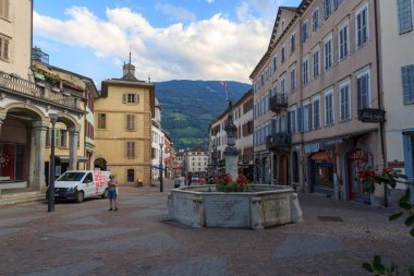 Sion, İsviçre - 11 Ağustos 2024: Eski Sion kasabasında arka planda dağ manzarası olan yaya bölgesi, dükkan ve çeşme. Sion, İsviçre 'nin Valais kantonunun merkezi kent..