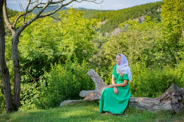 on a felled tree sits in a green dress a girl with a handkerchief on her head. High quality photo