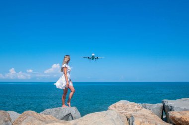 Airborne Encounters: Girl in White Dress on Stones, Plane Over the Blue Sea. High quality photo