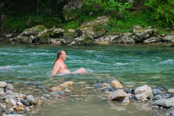 A young man with long hair is seen swimming in a crystal clear mountain river. The lush green trees and large rocks provide a scenic background, creating a peaceful and refreshing atmosphere.
