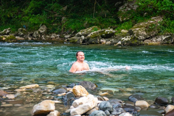Young man with long hair finding serenity while swimming in a pristine mountain river surrounded by lush trees and rocks, enjoying a moment of blissful tranquility in nature.