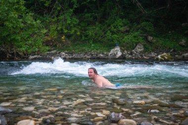 Dağ nehrinde yüzen uzun saçlı genç bir adamın sakin ve huzurlu bir sahnesi. Etrafı yosun kaplı kayalarla ve yemyeşil ağaçlarla çevrili doğanın güzelliğinde teselli bulur..