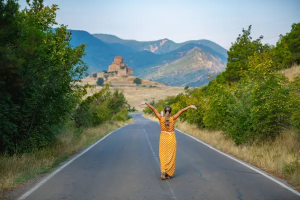A peaceful scene of a solitary female figure in an orange dress walking on a rural path, surrounded by lush greenery, heading towards a distant mountain and church on a sunny day
