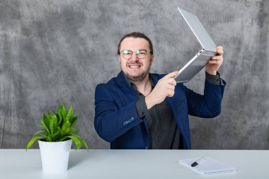 Mavi takım elbiseli genç bir profesyonel modern bir ofiste masa başında oturuyor ve dizüstü bilgisayarda yazıyor. Ofis parlak ve arka planda yemyeşil bir bitki var..