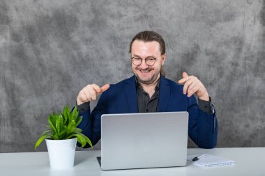 Mavi takım elbiseli ve gözlüklü profesyonel bir adam masada oturuyor ve bir dizüstü bilgisayar üzerinde çalışıyor. Arka planda verimli ve barışçıl bir çalışma ortamı yaratan yemyeşil bir bitki var..