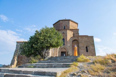Turuncu elbisesi ve beyaz atkısı olan sakin ve zarif genç bir kadın güneşli bir günde, görkemli bir tarihi kilisenin önünde zarif bir şekilde duruyor, sonsuz güzellik ve sükunet yayıyor..