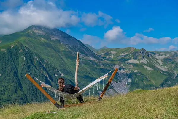 Tranquil Retreat: A man dressed in black apparel is peacefully resting in a hammock set against the backdrop of a lush mountain and azure sky scenery.