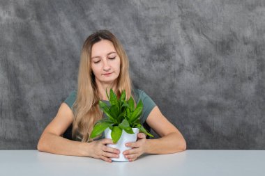 Uzun, dalgalı sarı saçlı genç bir kız ahşap bir masada oturuyor, tenceredeki parlak yeşil bir çiçeğe dikkatlice bakıyor, doğasına ve güzelliğine hayranlık ve hayranlık gösteriyor..
