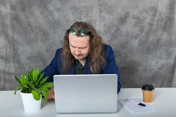 A young man with long hair is sitting at a clean and modern desk, working on a laptop computer with focus and determination in a contemporary workspace environment