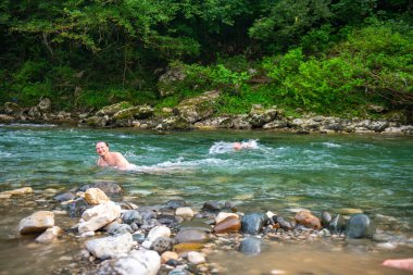 Dağlarda sakin bir nehir sahnesi, ağaçlar ve kayalar arasında yüzen uzun saçlı bir adam, huzurlu ve huzurlu bir doğal ortam yaratıyor..