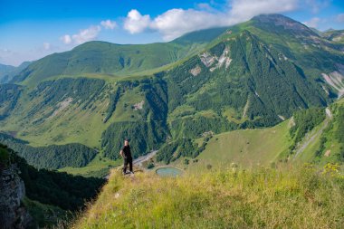 Koyu renk elbiseli yalnız genç bir adam soluk kesici, geniş yeşil dağların önünde duruyor, temiz mavi gökyüzü tarafından çerçevelenmiş, özgürlük ve macera hissi uyandırıyor..