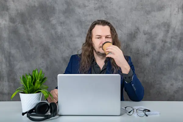 Stylish young man with long hair is confidently posing in a fashionable blue suit, holding a laptop and wearing headphones, exuding a trendy and modern vibe.